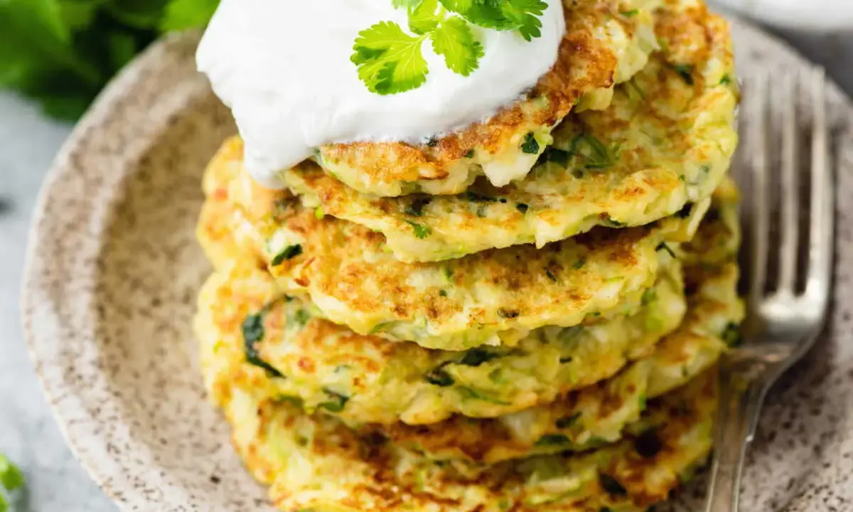 Pile de galettes de courgette dorées, garnies de crème et de coriandre fraîche, sur une assiette en céramique.