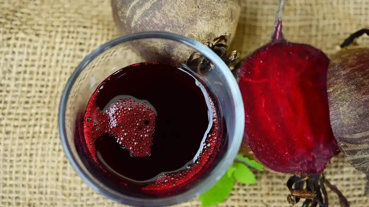 Verre de jus de betterave rouge vif, avec des betteraves entières et coupées autour. Une boisson saine pour savoir comment faire du jus de betterave.