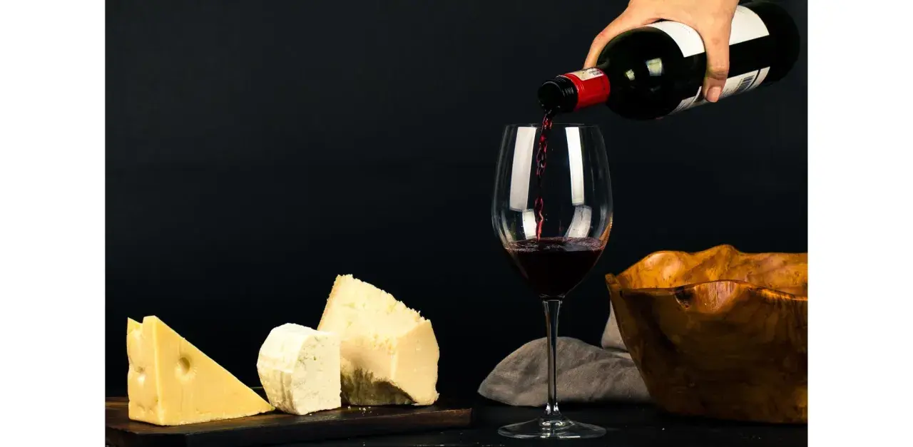Vin rouge versé dans un verre, à côté de morceaux de fromage. La question "quoi boire avec une raclette" trouve ici une réponse évidente.