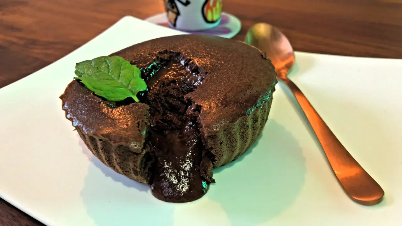 Un délicieux mi cuit chocolat coulant, garni d'une feuille de menthe, sur une assiette blanche. Une cuillère dorée est à côté.