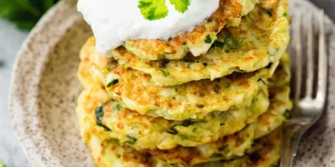 Pile de galettes de courgette dorées, garnies de crème et de coriandre fraîche, sur une assiette en céramique.
