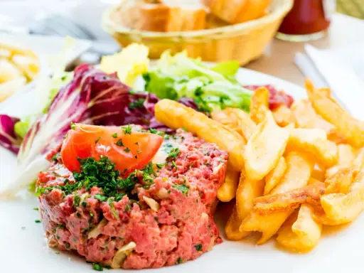 Un délicieux tartare de boeuf, garni de persil et d'une tranche de tomate, accompagné de frites dorées et d'une salade fraîche.