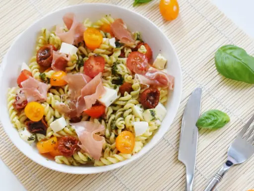 Une salade de pâtes originale avec des tomates cerises, du prosciutto, des dés de mozzarella et une sauce verte savoureuse.