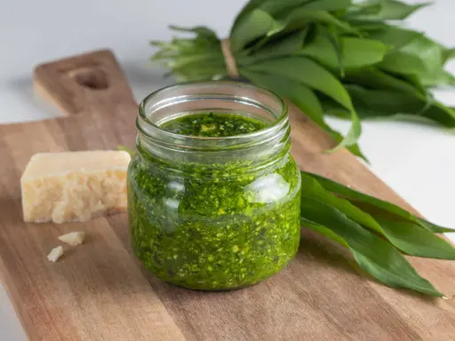 Pesto ail des ours fait maison dans un bocal, avec du fromage et des feuilles fraîches sur une planche en bois.