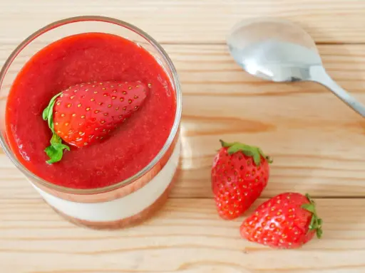 Un verre de panna cotta avec une fraise entière sur un coulis de fraise. Deux fraises fraîches et une cuillère à côté.