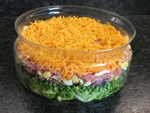 Une salade 7 étages colorée dans un grand bol en verre, avec des couches de laitue, petits pois, oignons rouges, jambon, œufs durs et fromage râpé.