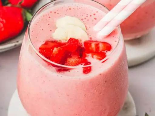 Un délicieux smoothie fraise banane, garni de morceaux de fruits frais et de pailles rayées roses et blanches.