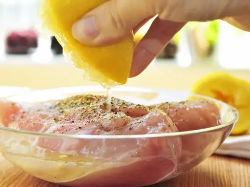 Préparation de la meilleure marinade poulet : un citron est pressé sur des morceaux de poulet assaisonnés dans un bol en verre.