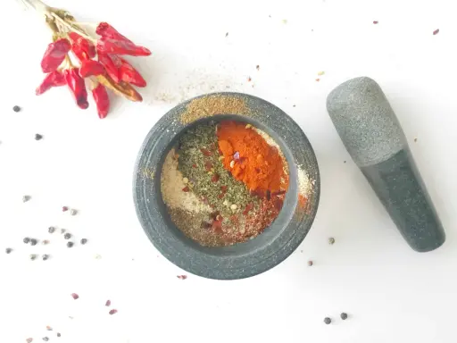 Mortier en pierre rempli d'épices pour fajitas, avec des piments rouges séchés et des grains de poivre à proximité.