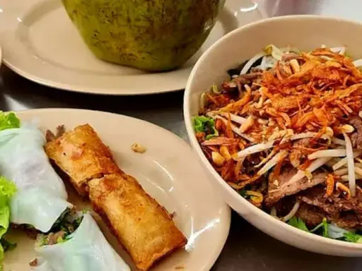 Un bol de bo bun garni de viande, légumes et oignons frits, accompagné de rouleaux de printemps et de sa sauce.