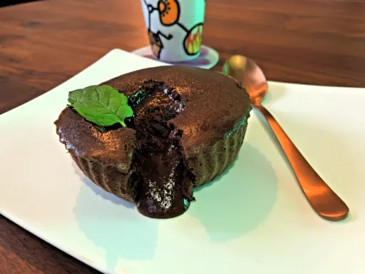 Un délicieux mi cuit chocolat coulant, garni d'une feuille de menthe, sur une assiette blanche. Une cuillère dorée est à côté.