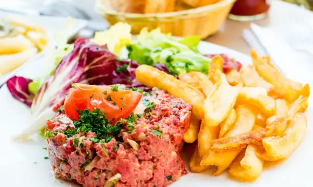 Un délicieux tartare de boeuf, garni de persil et d'une tranche de tomate, accompagné de frites dorées et d'une salade fraîche.