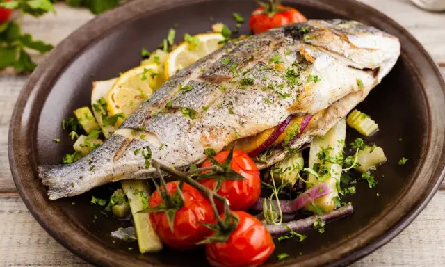 Poisson entier, probablement une truite au barbecue, servi avec des tomates cerises, des quartiers de citron et des herbes fraîches sur une assiette marron.