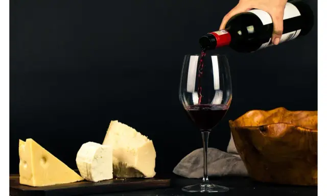 Vin rouge versé dans un verre, à côté de morceaux de fromage. La question "quoi boire avec une raclette" trouve ici une réponse évidente.