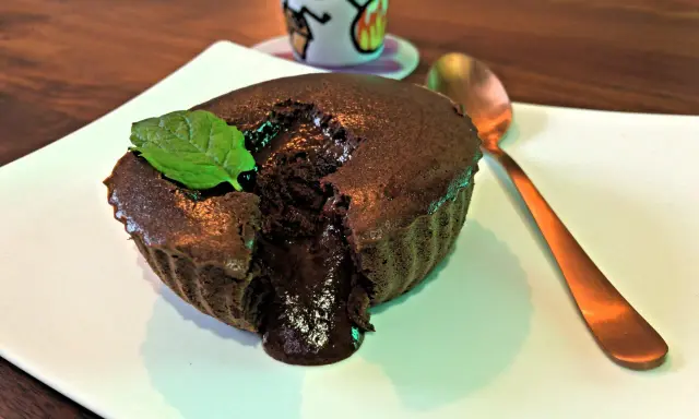 Un délicieux mi cuit chocolat coulant, garni d'une feuille de menthe, sur une assiette blanche. Une cuillère dorée est à côté.