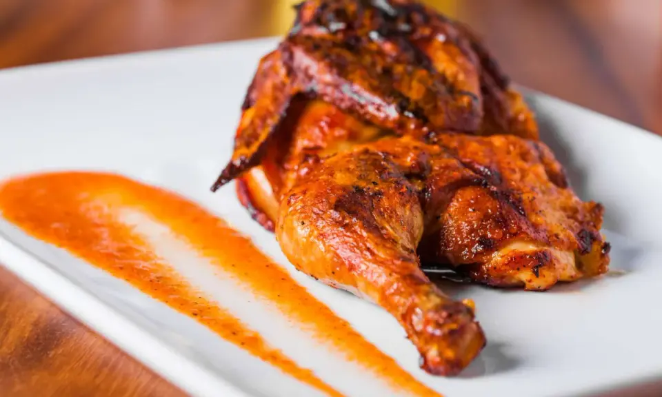 Un délicieux poulet portugais au four, la peau dorée et croustillante, servi avec une sauce épicée sur une assiette blanche.