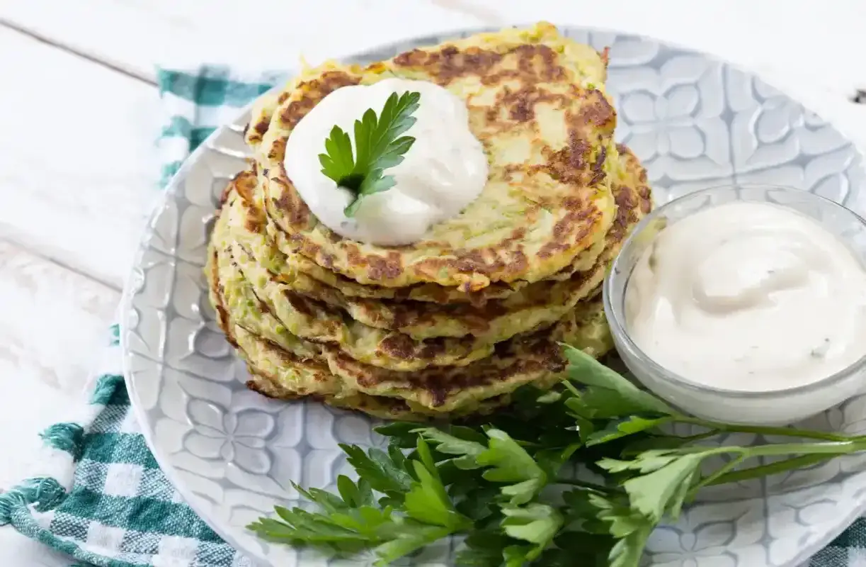 Pile de galettes de courgette dor&eacute;es, garnies d'une cuill&egrave;re de cr&egrave;me et de persil frais, servies avec une sauce cr&eacute;meuse.
